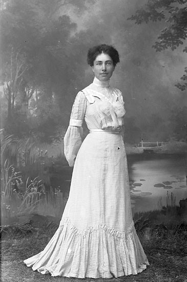 Svartvitt Foto. En kvinna i 30-årsåldern står upp framför en fotovägg föreställande en näckrosdamm. Hon bär en randig plisserad kjol och skjorta från tidigt 1900-tal. 