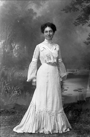 Svartvitt foto. En leende kvinna i 30-årsåldern står upp framför en fotovägg som föreställer en näckrosdamm. Hennes skjorta och kjol, från tidigt 1900-tal, är i ett randigt plisserat mönster med vita band. 