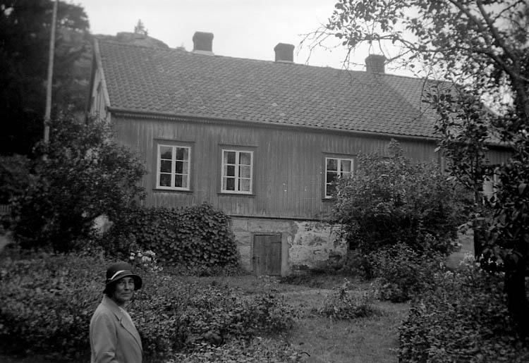 Svartvitt foto. Kvinna i 40-årsåldern i hatt från 1920-tal står i en trädgård omringad av träd och buskar. I bakgrunden finns ett bohuslänskt hus med pröjsade fönster.