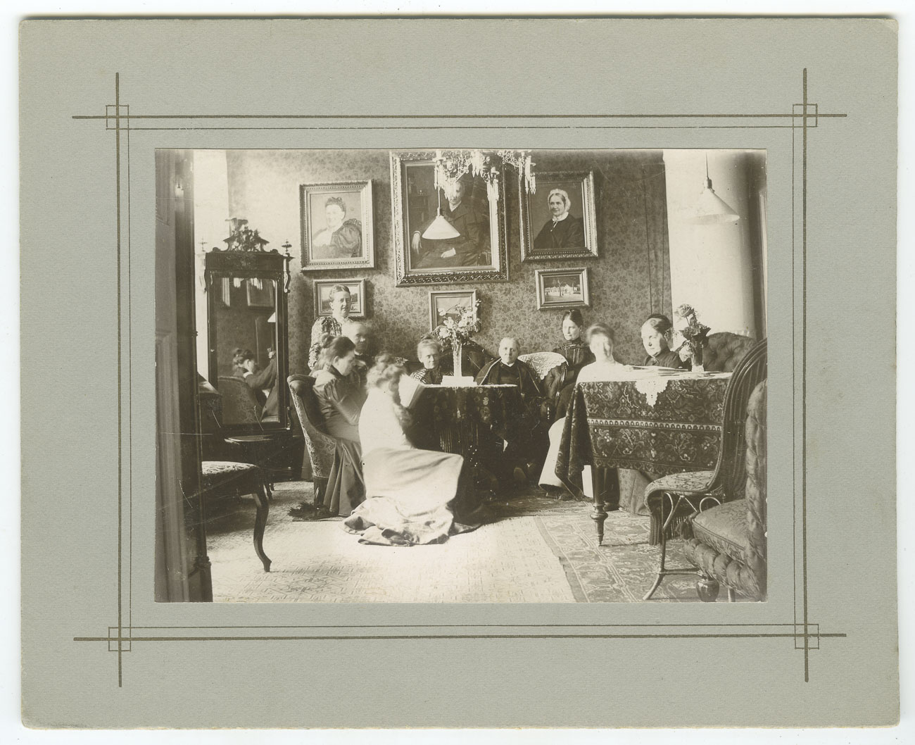 Svartvitt foto. nio kvinnor sitter och står i ett vardagsrum från tidigt 1900-tal. Möblerna är mörka och på väggen bakom kvinnorna hänger sex målade tavlor i guldramar. 