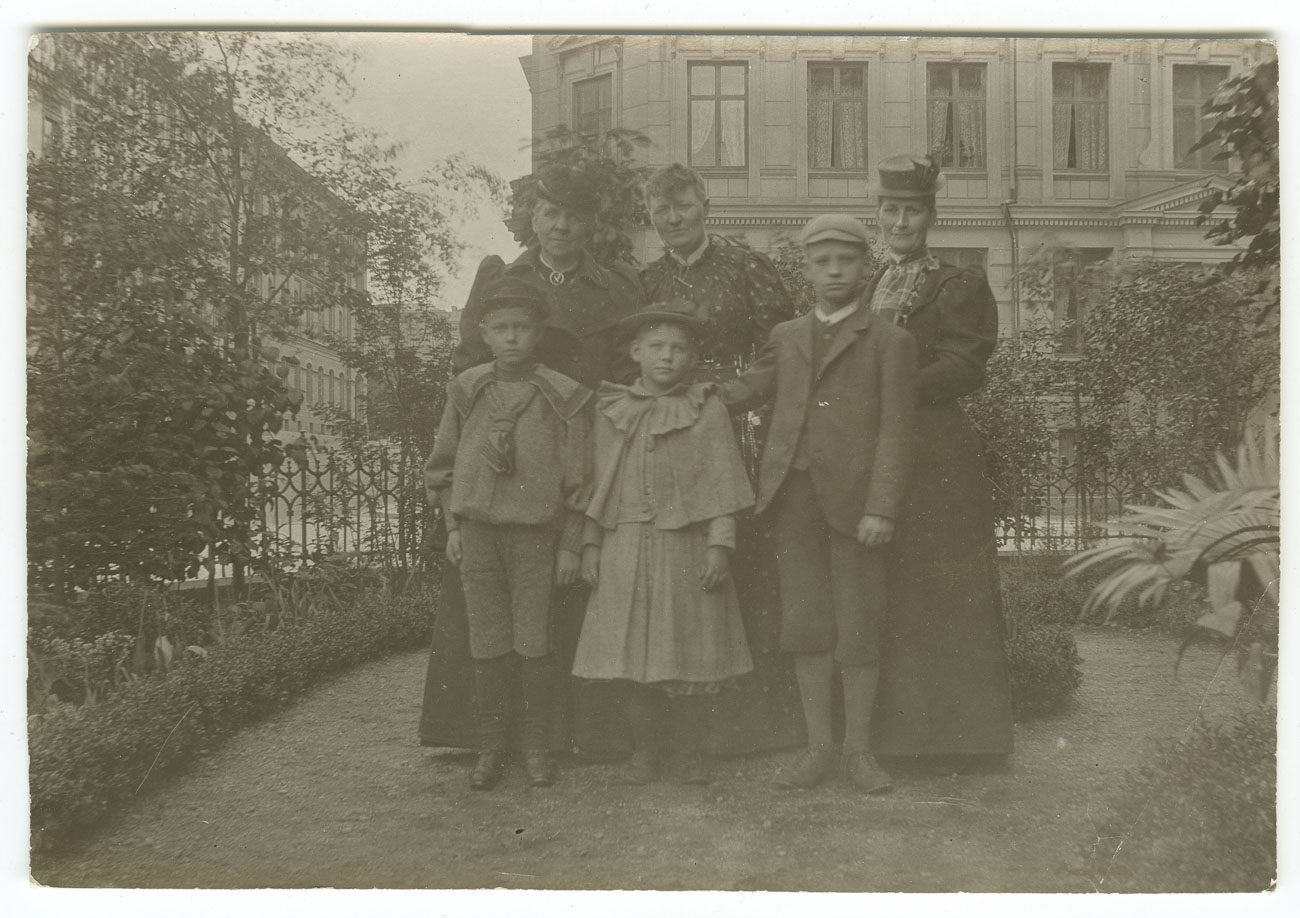 Svartvitt foto. sex personer, tre kvinnor och tre barn, står i en stadsträdgård. Kvinnan i mitten har kortklippt hår.