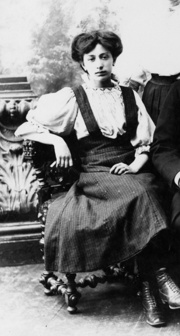 Svartvitt foto. Kvinna i övre tonåren sitter på en stol. Hon har en vit skjorta med stora puffärmar och en mörkmönstrad hängselkjol från tidigt 1900-tal. Hennes mörka hår är uppsatt i en lockig frisyr.