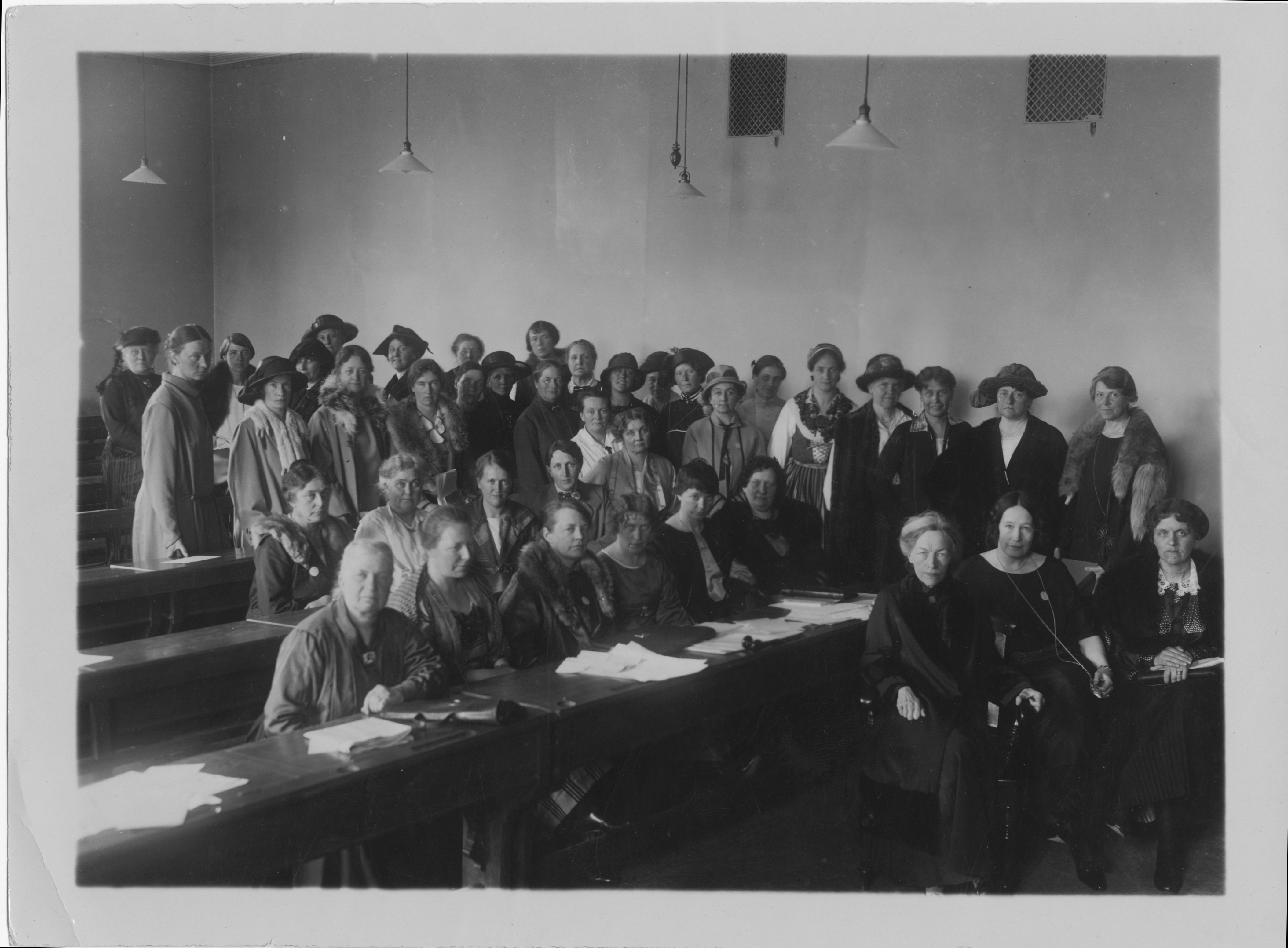 Svartvitt foto. Gruppbild med 41 kvinnor i olika ålder. De befinner sig i en skolsal. På bänkraderna finns det vita pappersark utspridda och en ordförandeklubba. 