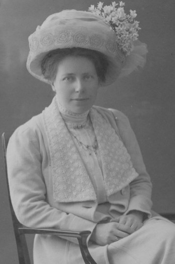 Svartvitt foto. Kvinna i 30 års åldern sitter i en stol. Hon har på sig en ljus kappa med en vit plisserad blus. Hennes mörka hår är uppsatt under en ljus hatt från tidigt 1900-tal. Hatten är prydd med vitsippor och vitt flor.