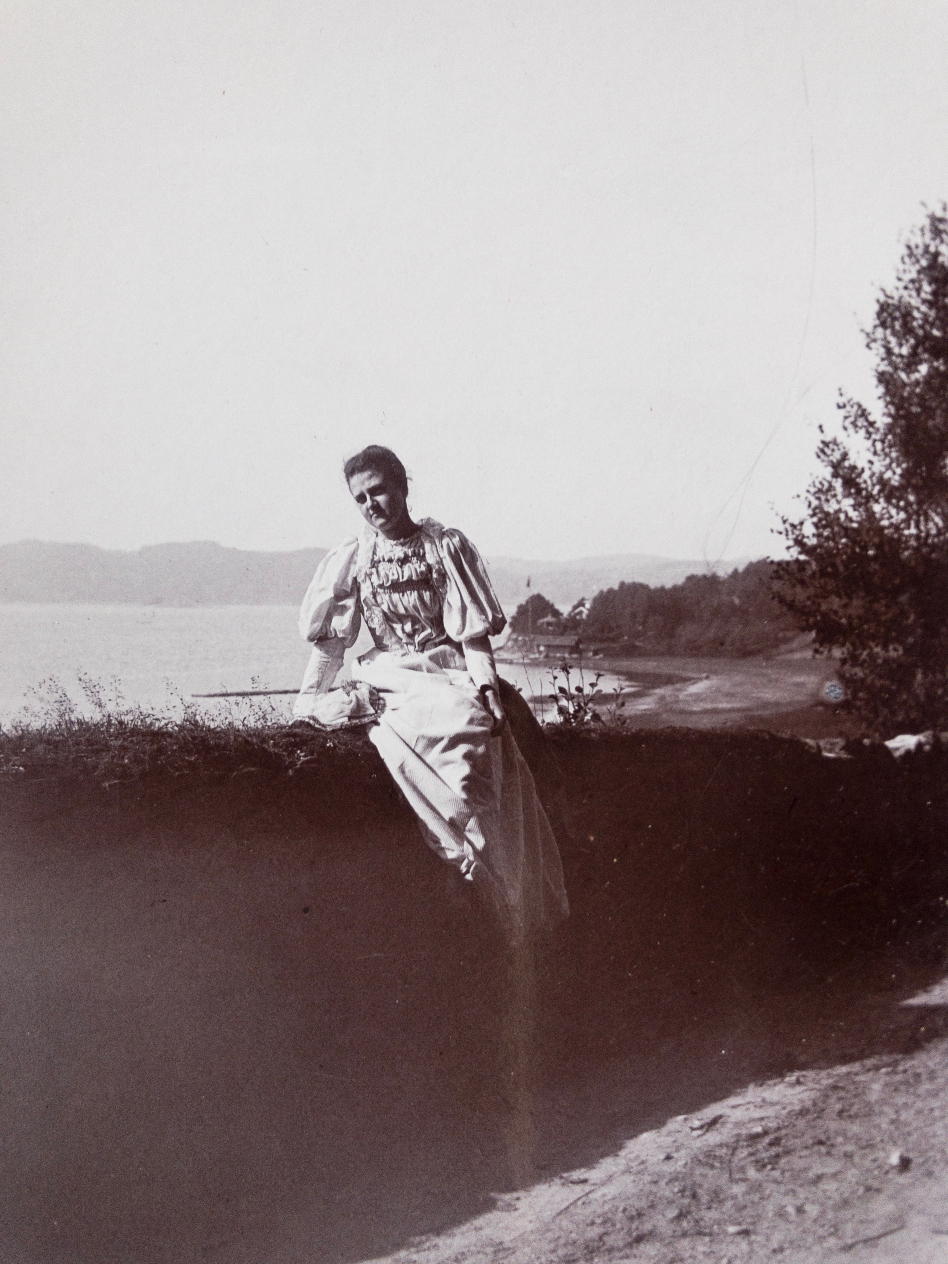 Svartvitt foto. Kvinna i 20 årsåldern sitter på en vall. Bakom henne syns havet och ett hus. Hon har på sig en ljus plisserad klänning med puffärm. 