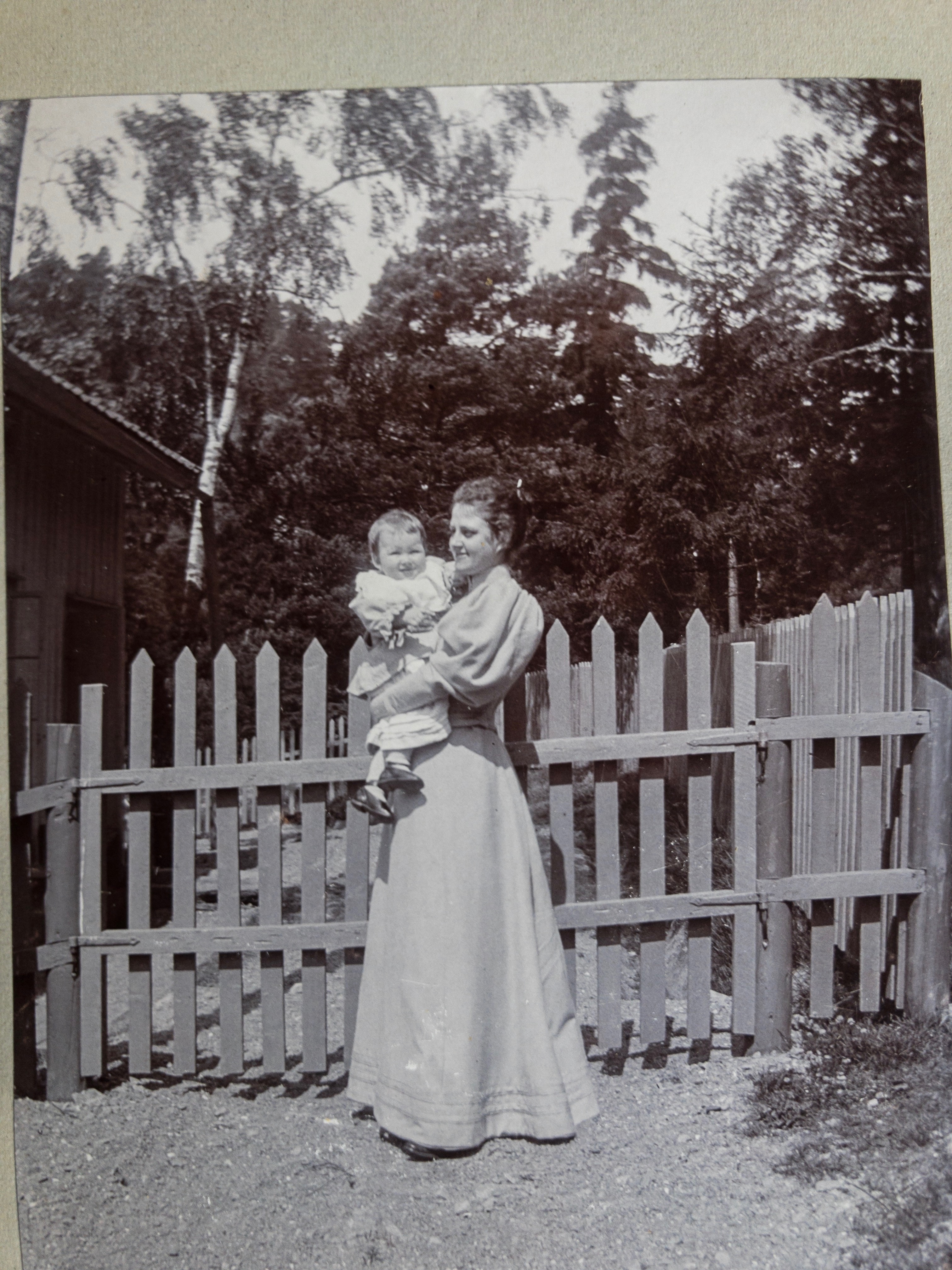 Svartvitt foto. Kvinna i 30 års åldern som håller ett barn i två års ålder i sin famn. De står framför en grind med träd och buskar bakom.