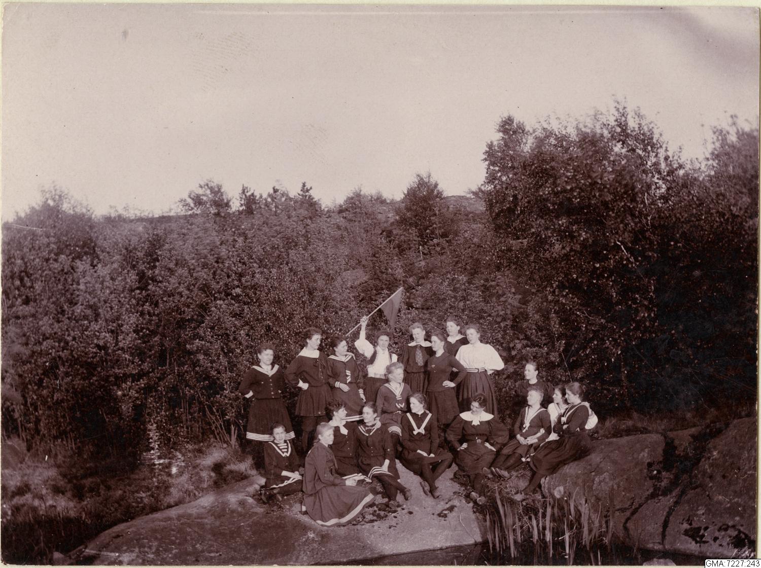 Svartvitt foto. Gruppbild med 18 kvinnor som står på en bohuslänsk klippa med träd runt sig. Kvinnorna bär gymnastikkläder. Kvinnan ståendes i mitten viftar med en flagga.