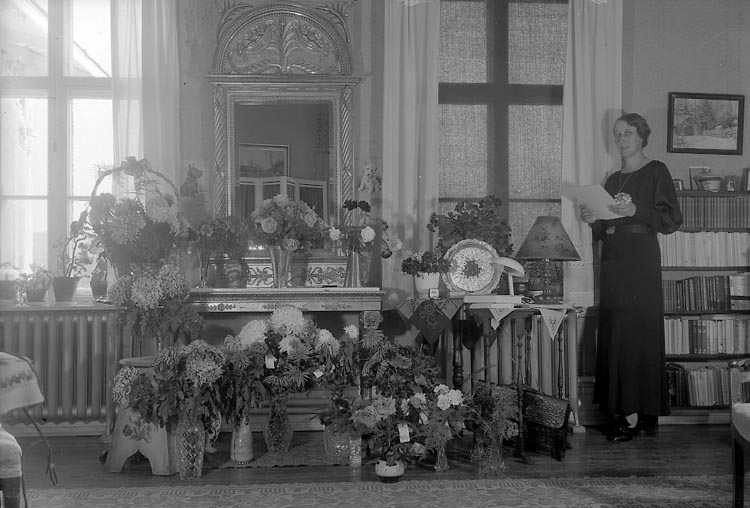 Svartvitt foto. Kvinna 50 år. Hon står i höger i bild bredvid ett 20-tal buketter och blomsterkorgar. I hennes händer håller hon en bunt med telegram.